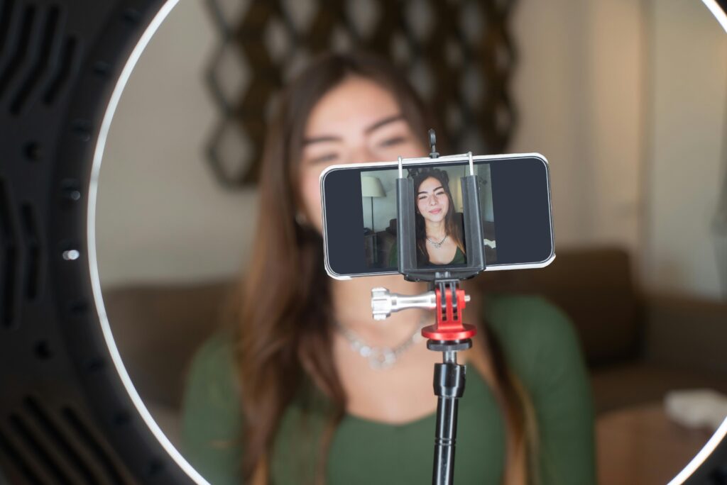 A young woman live streams from home using a smartphone and ring light setup.